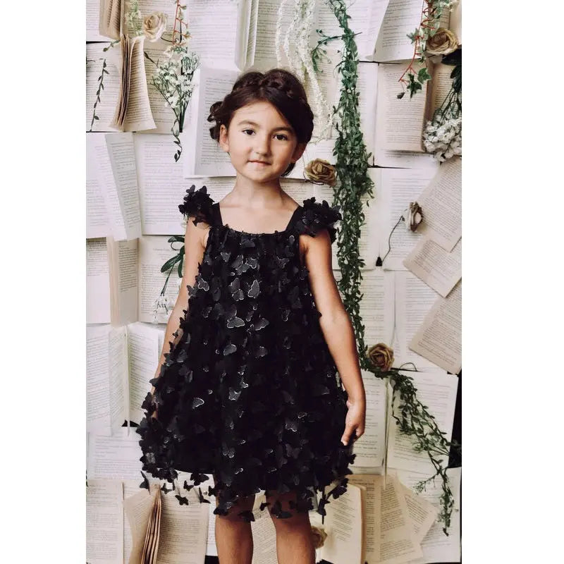 Dolly by Le Petit Tom Allover Butterflies Tutu Dress Black; a girl stands in a layered black tulle dress with butterfly appliques.