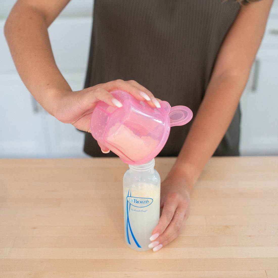 A person carefully pours baby formula from a pink Dr. Brown's Baby Formula Dispenser into a baby bottle on a wooden countertop. The transparent bottle with blue and white designs sits against the backdrop of a cozy white kitchen setting