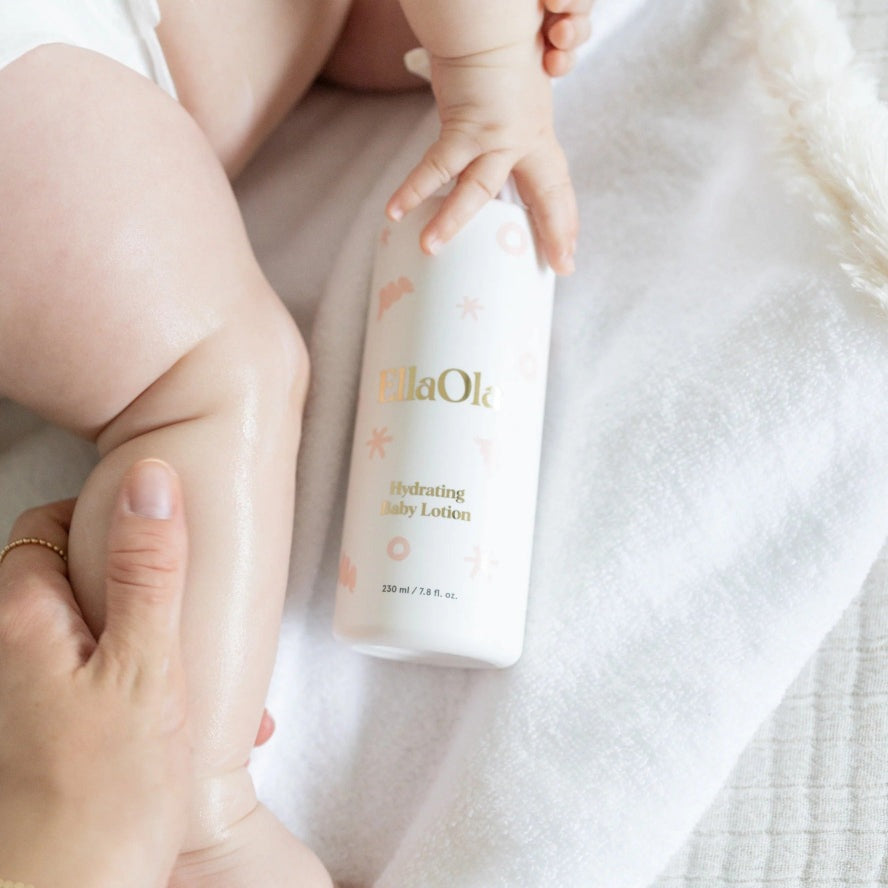 EllaOla Fragrance Free Hydrating Baby Lotion 230ml applied to a baby's leg by a caregiver on a soft white towel background.