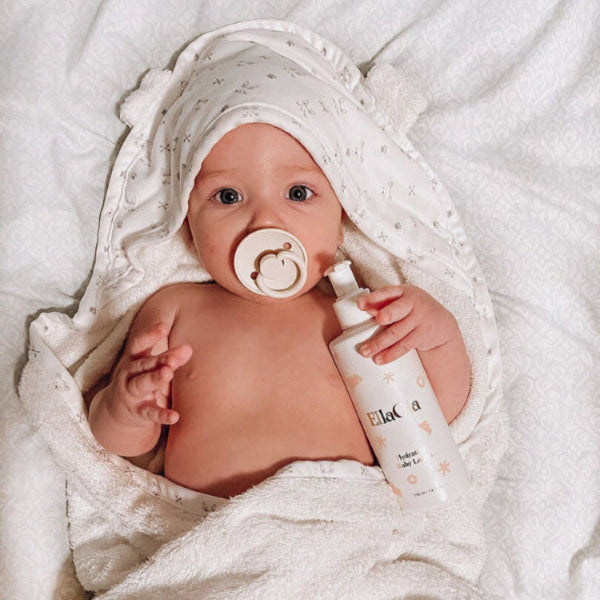 EllaOla Fragrance Free Hydrating Baby Lotion 230ml: baby swaddled in white towel holds the lotion bottle and pacifier.