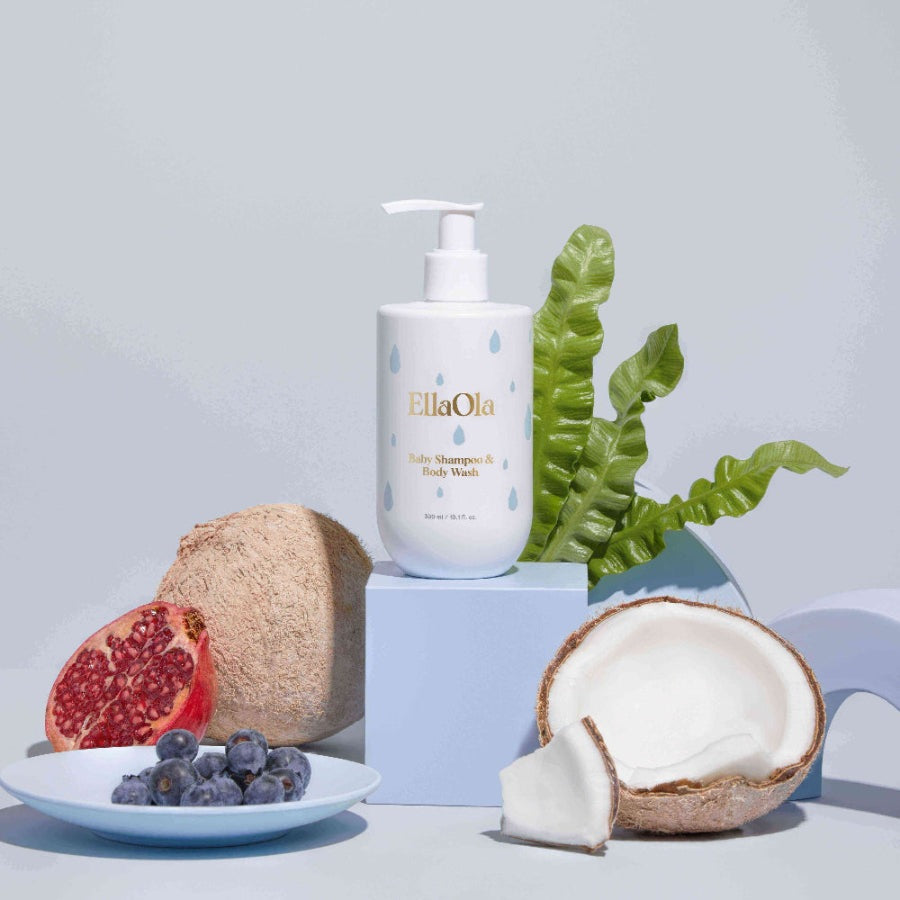 EllaOla Tear Superfood Baby Shampoo & Body Wash 300ml pump bottle sits on a pastel block amid fruit and leaves.
