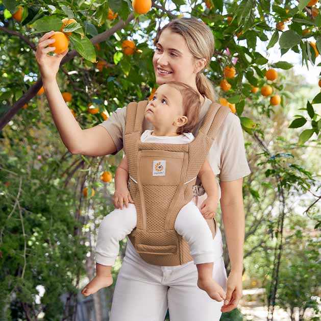 ERGOBABY Alta Hip Seat Baby Carrier in beige worn by mother and smiling baby reaching for oranges in a sunlit orchard.