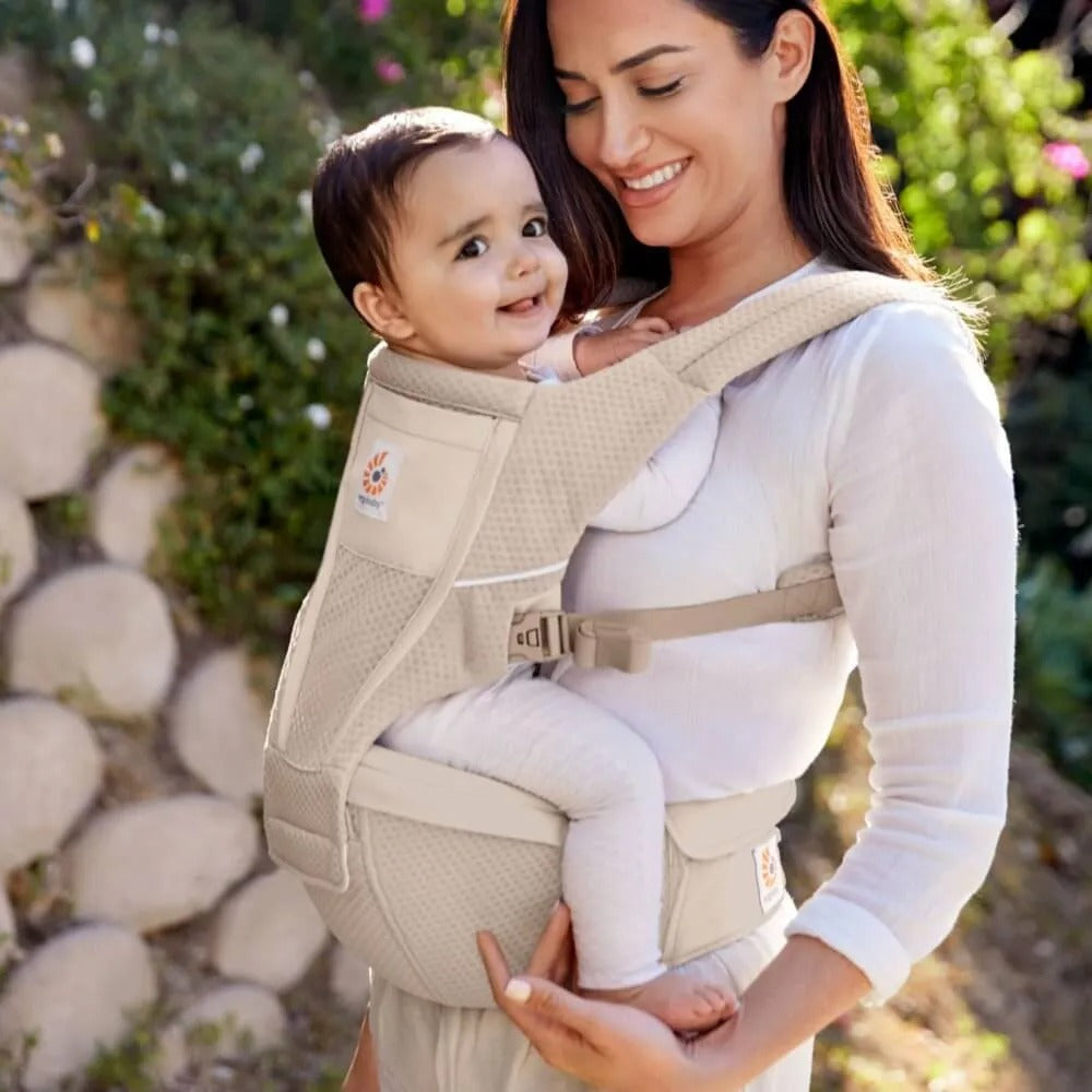 Ergobaby Alta Hip Seat Baby Carrier in beige, mom securely carries infant on her front hip with padded straps.