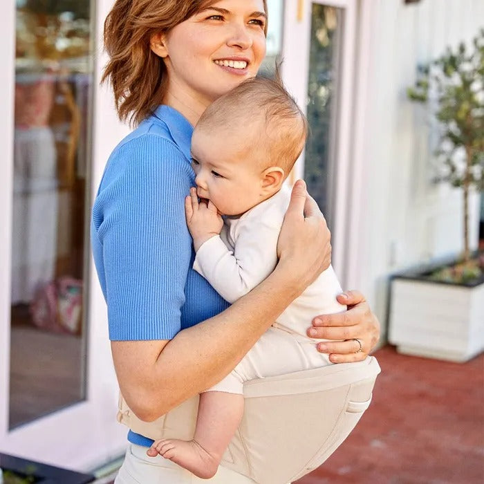 ERGOBABY Lift Hip Seat Baby Carrier Natural Beige worn by a smiling mom cradling a baby on her hip.