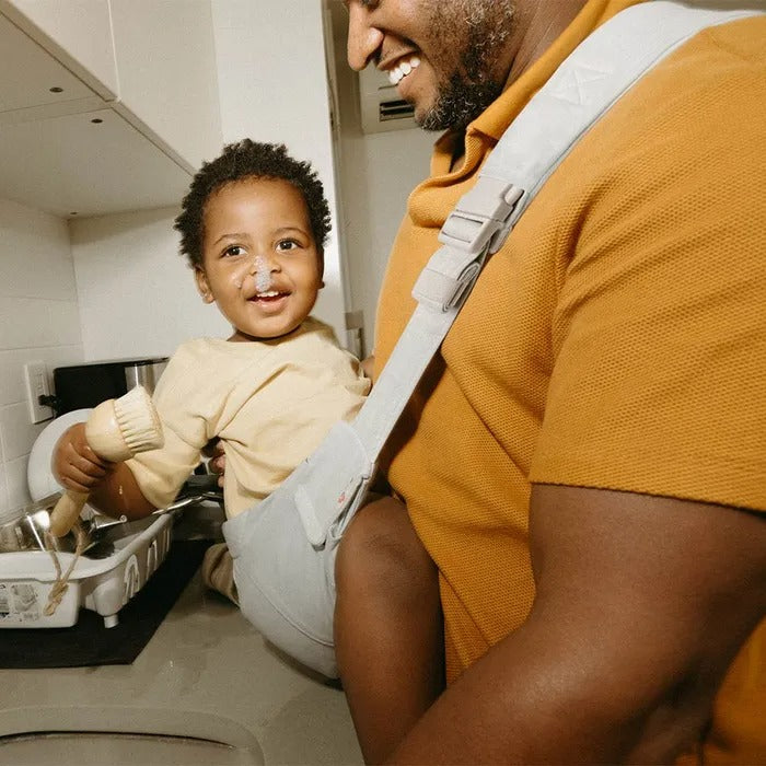 ERGOBABY Upsie Sling Carrier in light gray, worn by a smiling dad with toddler securely seated in it.