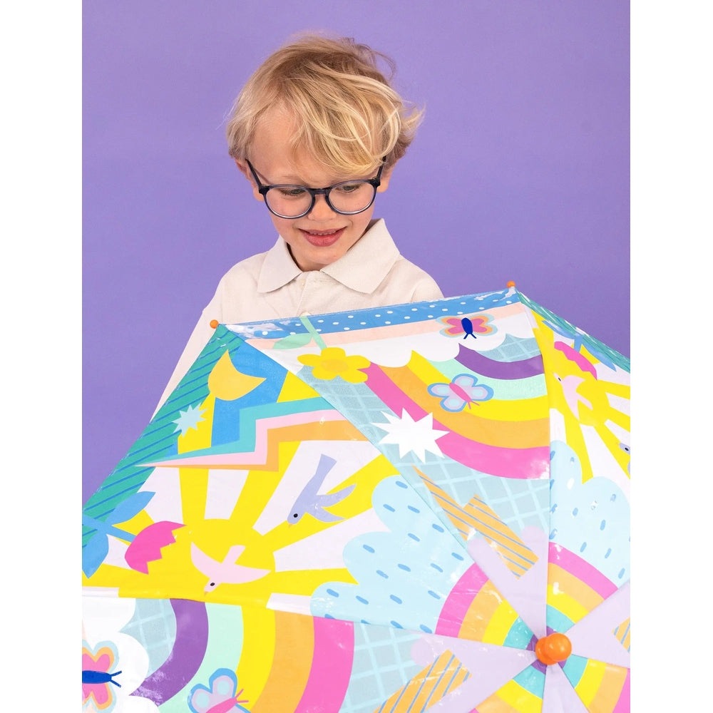 Floss And Rock Color Change Umbrellas Rainbows umbrella opened as a smiling child in a white shirt holds it.