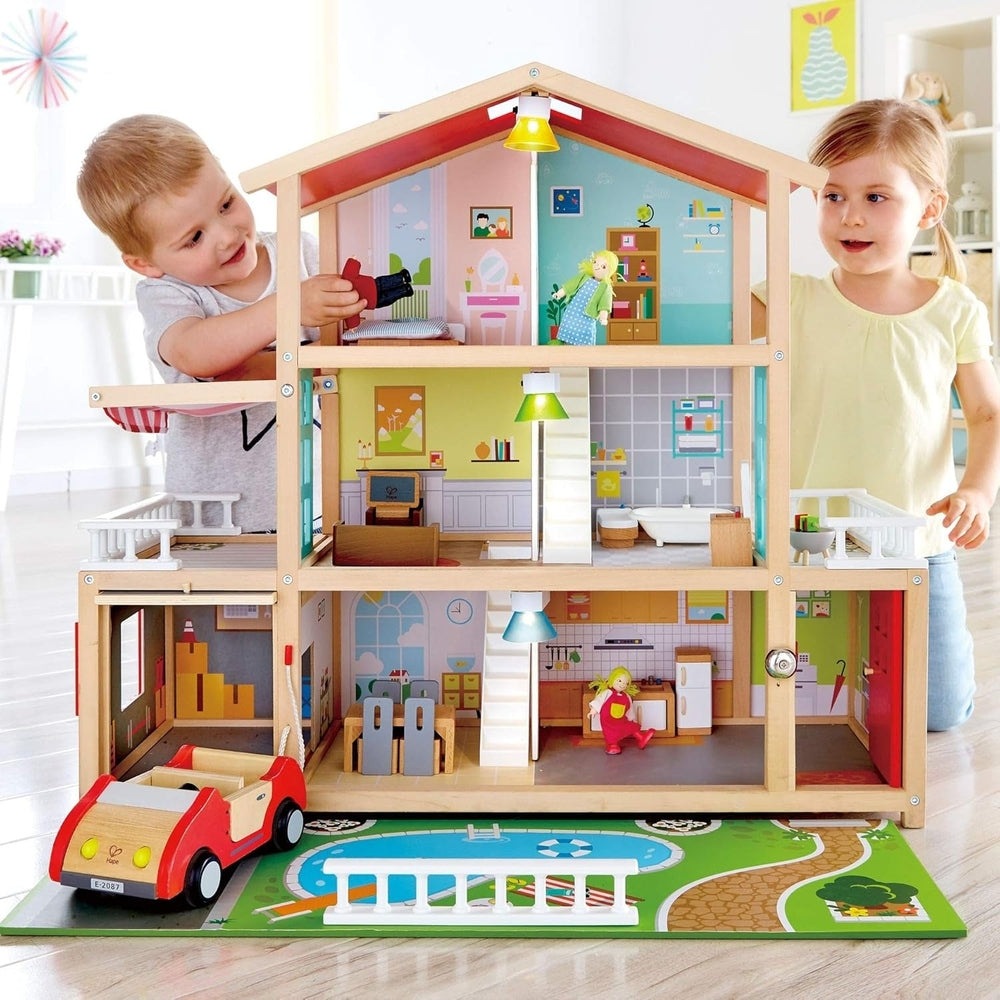 Hape Doll Family Mansion, wooden multi-level dollhouse with bright interiors; two kids play with tiny figures and a red car.