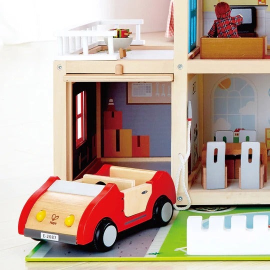 Hape Doll Family Mansion wooden dollhouse playset; a red wooden car exits the garage