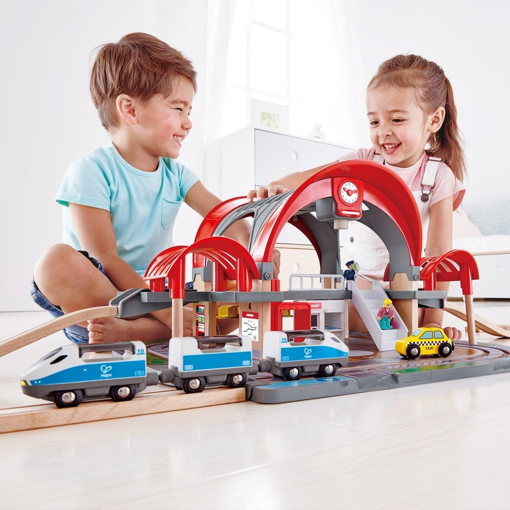 Hape Grand City Station train set with red arches and multi-level track; kids play with blue and white trains.