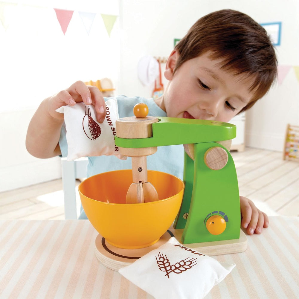 Hape Mighty Mixer wooden toy in green with yellow bowl, as a child pours flour from a bag into the mixer.