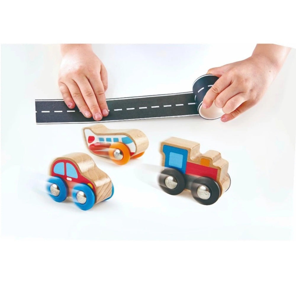 Hape Tape & Roll Train wooden toy set; hands lay and roll a dashed-road tape under colorful vehicles.