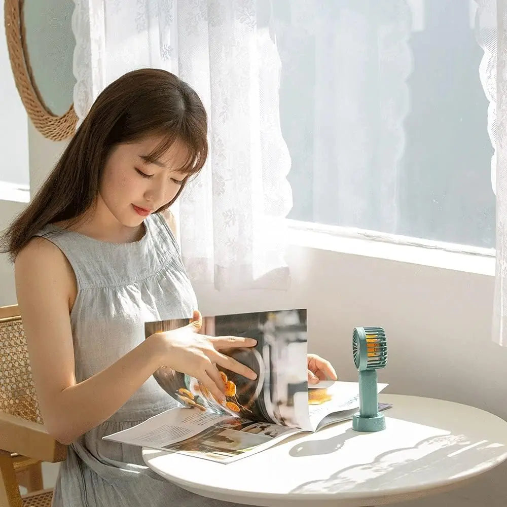 I2 Handheld 3-Speed Lithium Battery Fan Mini mint green on table beside magazines, ready for portable cooling.