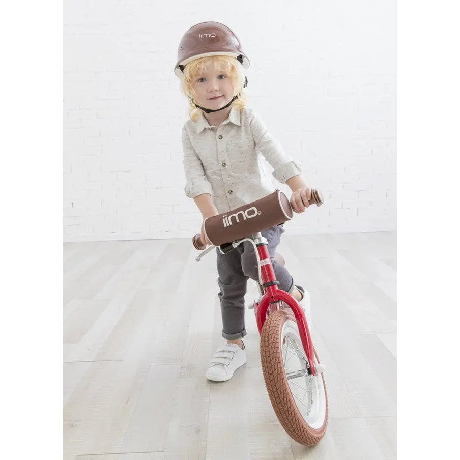 Iimo Balance Bike, red frame with brown padded handlebars; child rides indoors wearing helmet.