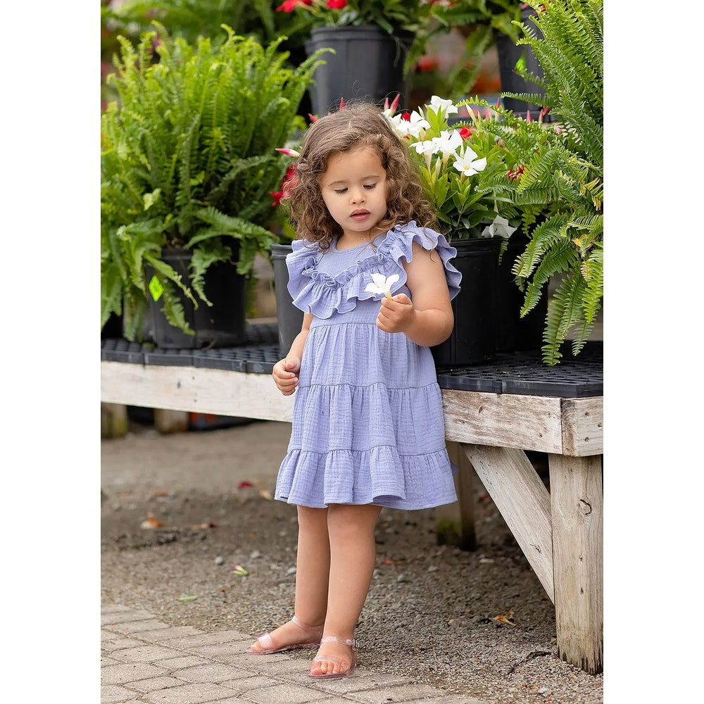 Isobella and Chloe Perry Pretty Cotton Gauze Dress Purple on a toddler girl, holding a flower amid potted ferns.