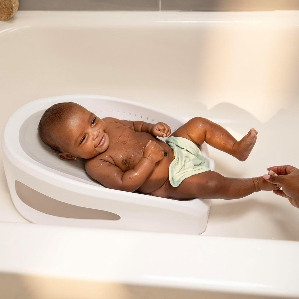 Ity by Ingenuity Easy Rinse Bather baby resting in a white plastic bath seat in the tub