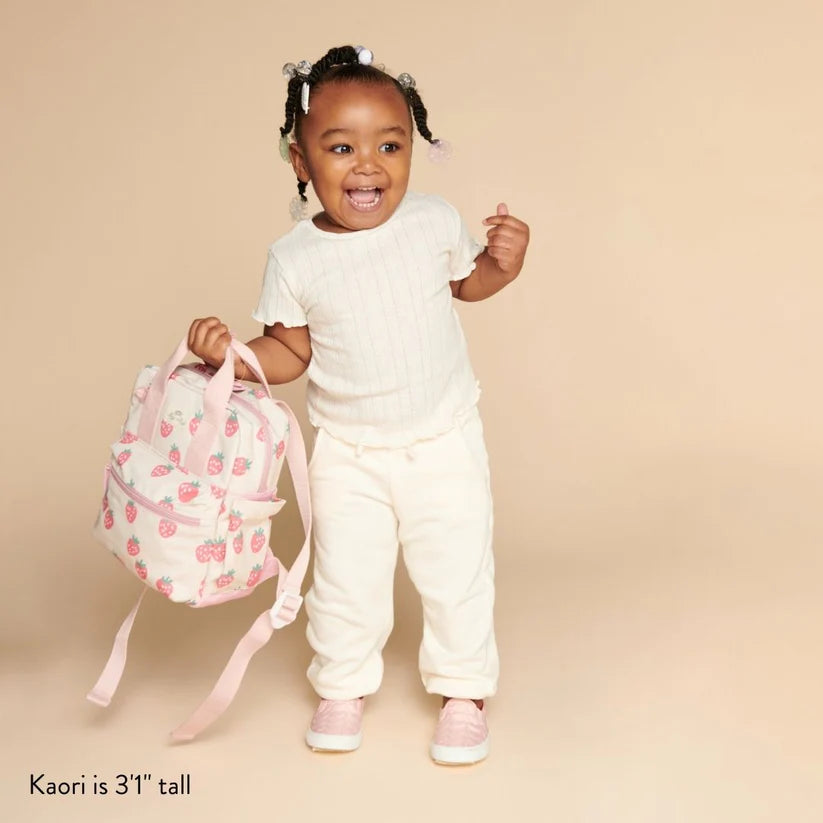 Itzy Ritzy Backpack in pink strawberry print held by a joyful toddler in a white outfit.