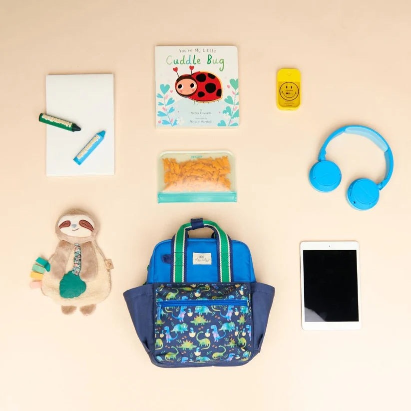 Itzy Ritzy Backpack blue body, dinosaur-pattern front pocket; green straps shown with kid-friendly school items.