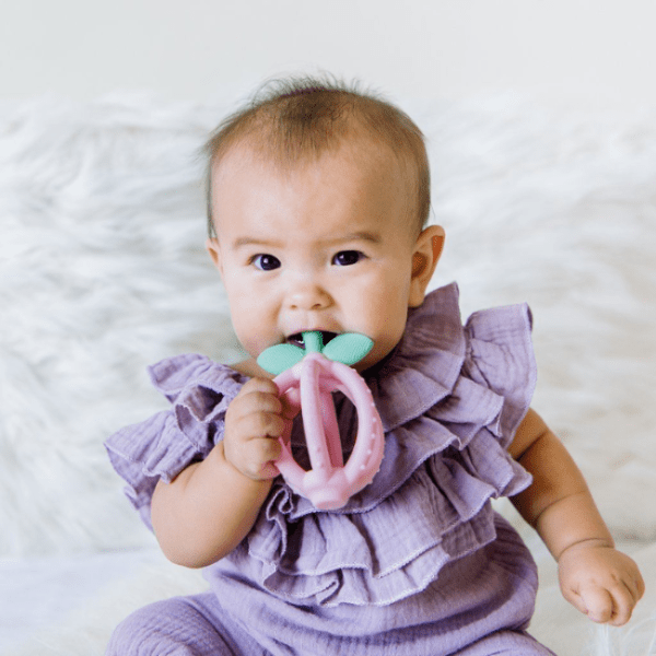 Itzy Ritzy Bitzy Biter Lemon Ball teether in pink with mint leaf, held by baby in purple outfit