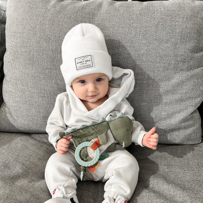 Itzy Ritzy Bitzy Crinkle Sensory Toy with Teether; teal ring teether worn by baby in white outfit on gray couch