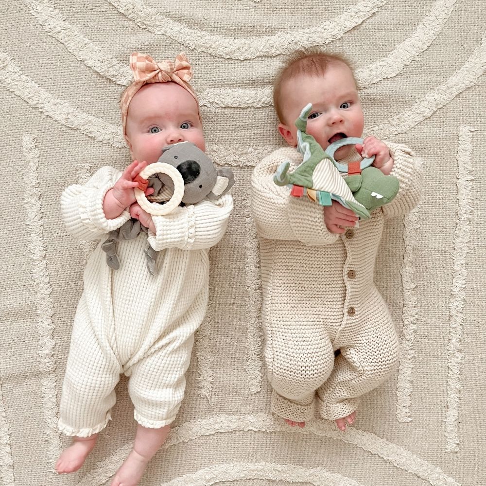 Itzy Ritzy Bitzy Crinkle sensory toy with teether; two babies lie on a cream rug holding plush teethers