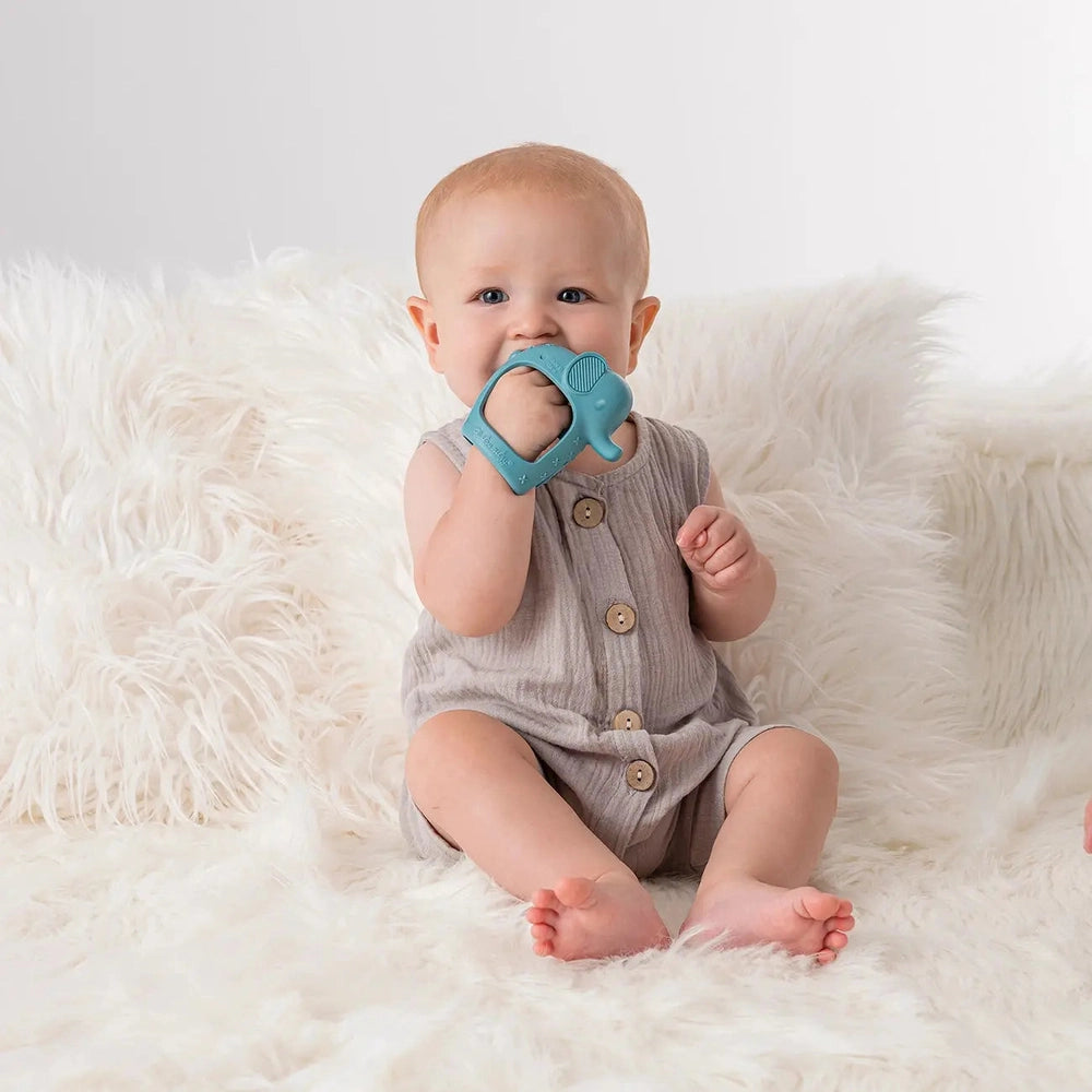 Itzy Ritzy Bitzy Grip teether in blue silicone, baby chews and holds it on a fluffy white backdrop.