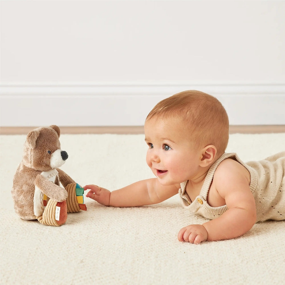 ITZY RITZY Bitzy Mirror Theo the Bear plush toy with colorful rings, as a baby reaches to grab them.