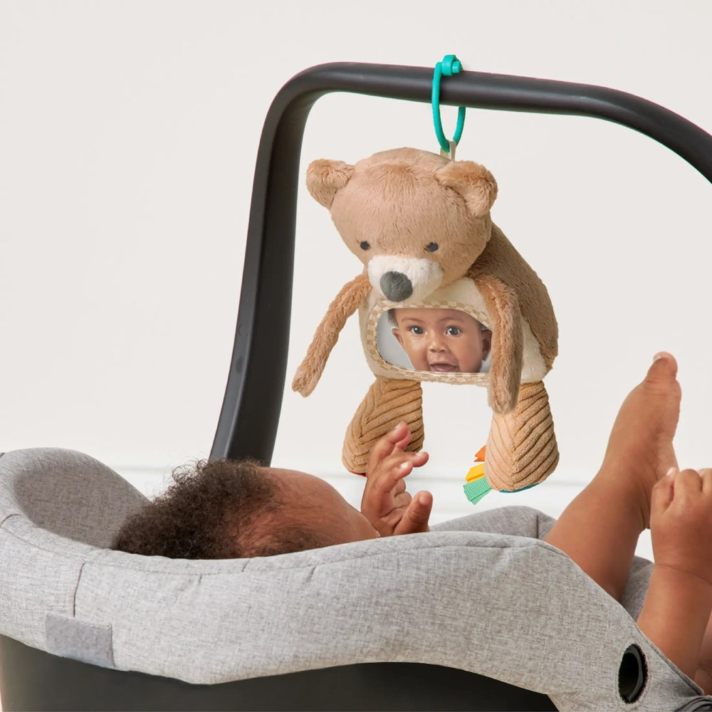 Itzy Ritzy Bitzy Mirror Theo the Bear hanging from a stroller arch, plush brown bear with a mirror window for baby to peek through.
