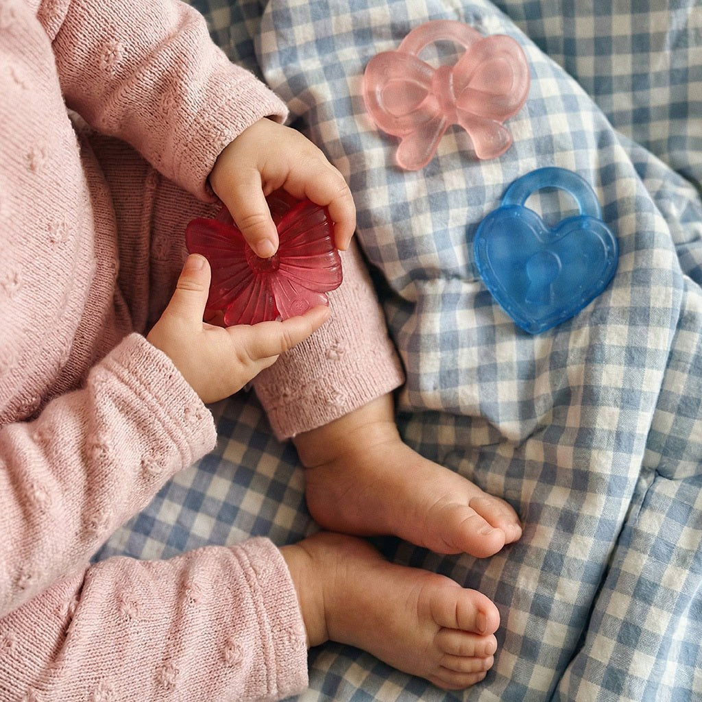 Child holding the deep pink bow teether beside the blue heart and light pink bow shapes