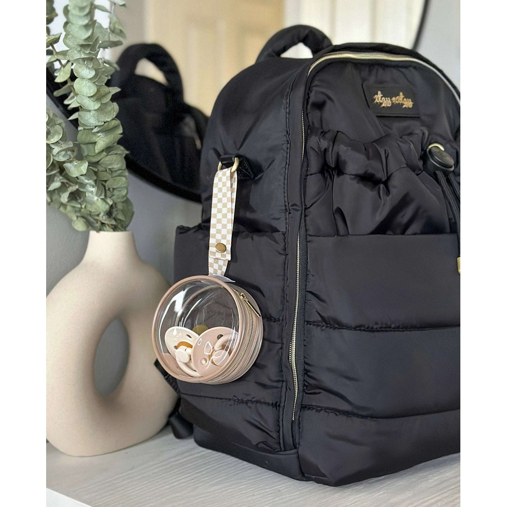 Itzy Ritzy paci case attached to a black puffer backpack with beige checkered strap and clear round pacifier holder