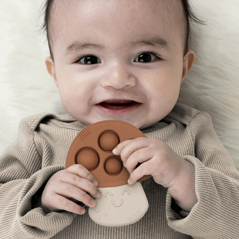 Itzy Ritzy Itzy Pop teether in tan silicone with cream handle held by a smiling baby
