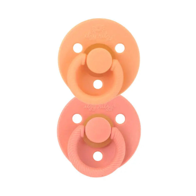 Itzy Ritzy Itzy Soother natural rubber pacifier set in peach and pink, two 0–6M pacifiers stacked with shields visible.