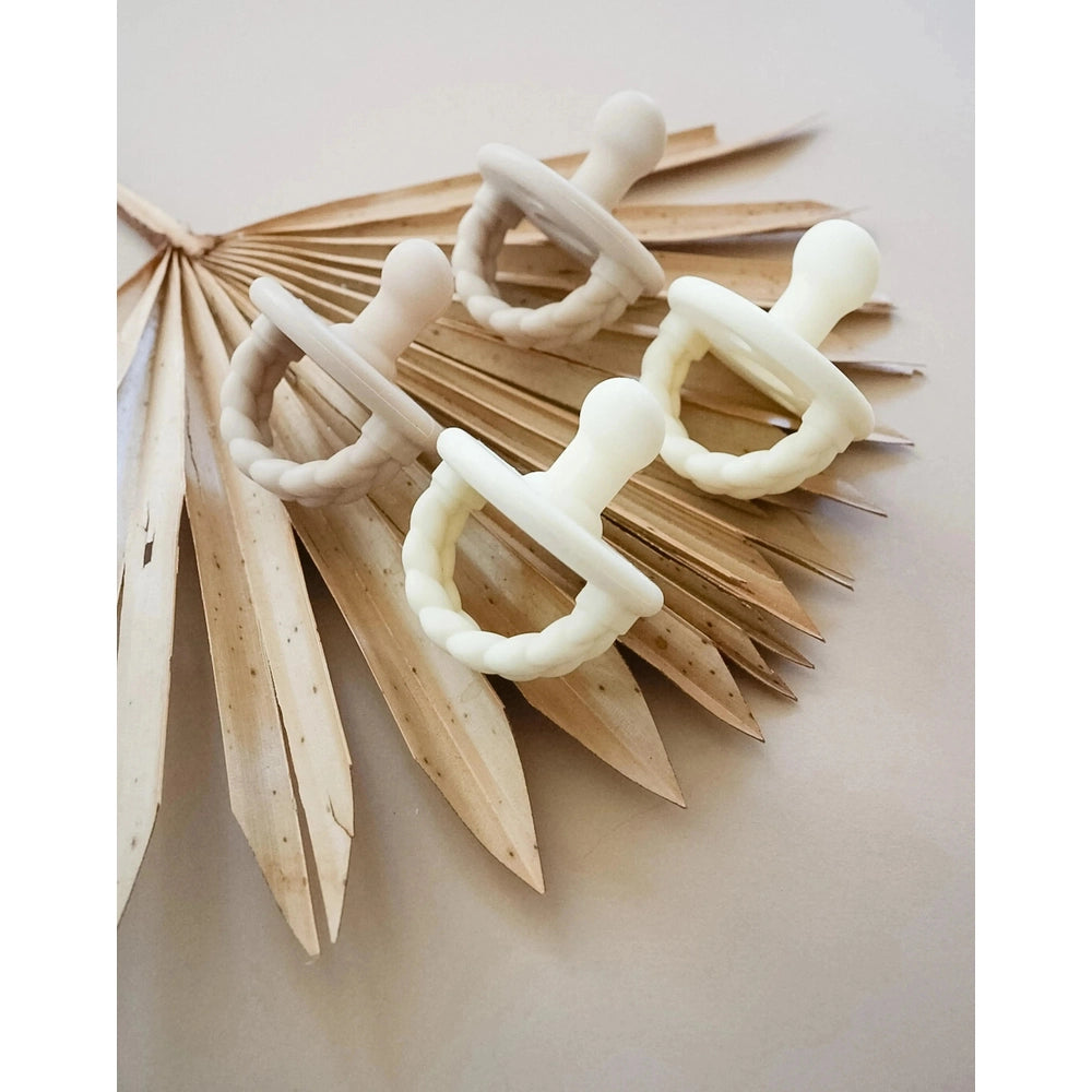 Itzy Ritzy Itzy Soother Natural Rubber Pacifier Sets (0–6M): four white pacifiers with braided handles shown on a dried palm leaf.
