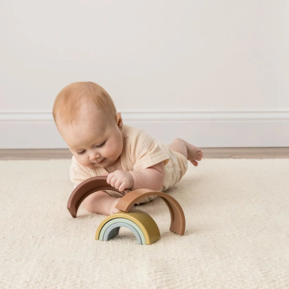 Itzy Ritzy Rainbow Stacking Toy; pastel wooden arches with a small rainbow, baby on a cream carpet.