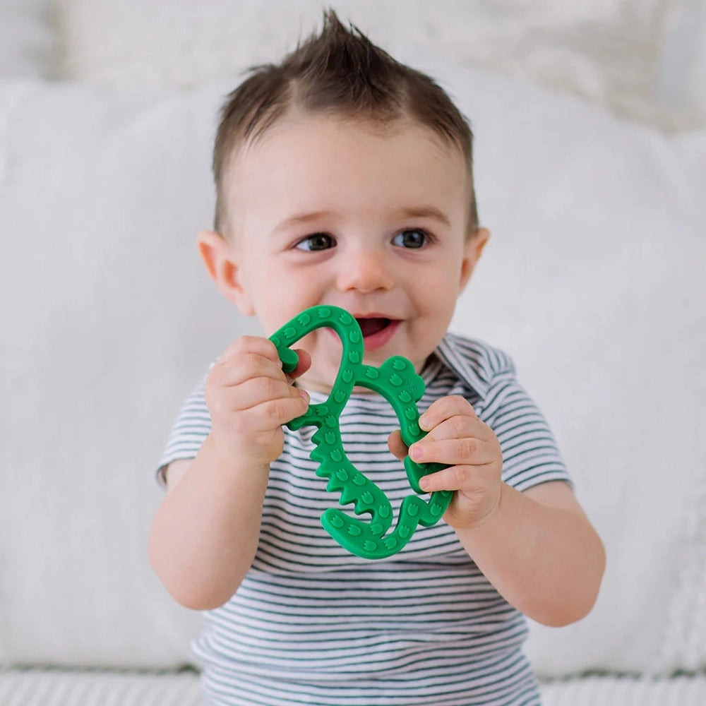 Itzy Ritzy silicone teether in green held by a smiling baby in a striped onesie