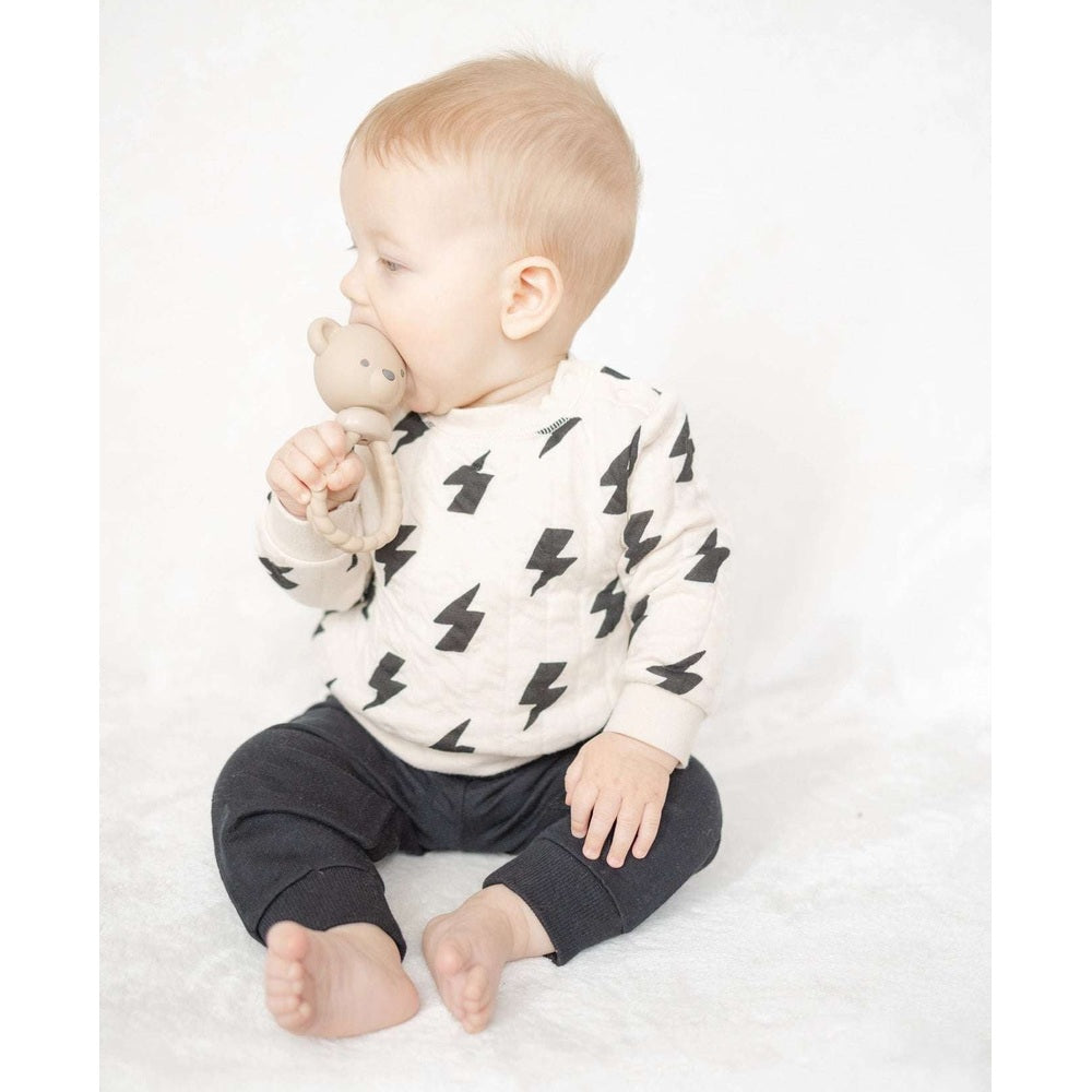 Itzy Ritzy Sweetie Rattle Bear teether in beige, held by a baby in a cream shirt with black bolt pattern