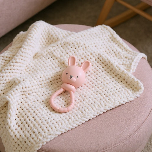 Itzy Ritzy Sweetie Rattle Pink Bunny resting on a cream knit blanket, highlighting the soft rattle with pink ring handle.