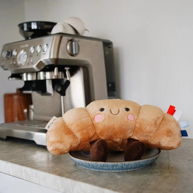 Itzy Ritzy Sweetie Snuggles Plush orange crab toy resting on a plate on a kitchen counter beside a coffee machine