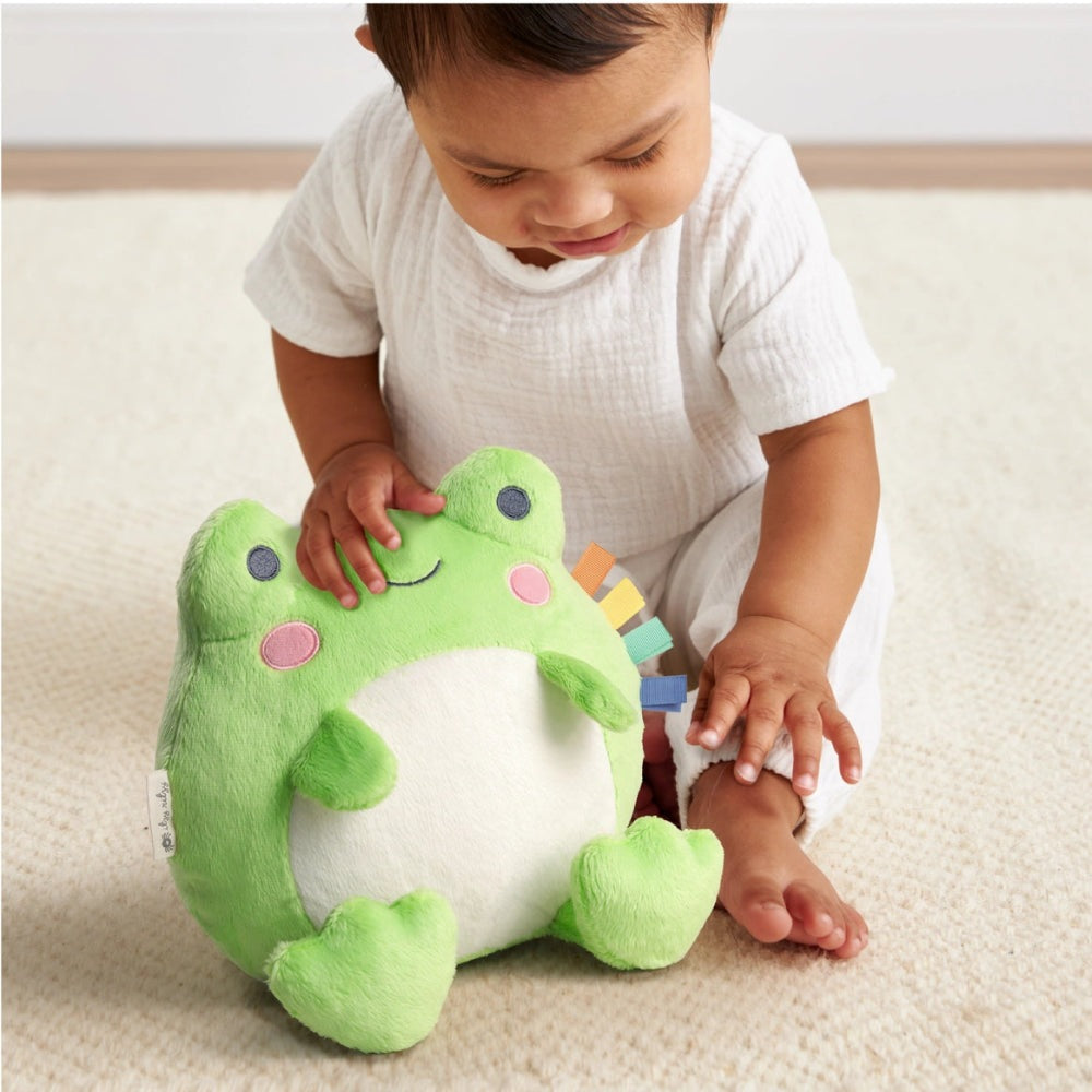 Itzy Ritzy Sweetie Snuggles Plush frog toy; green frog with multicolor tabs, toddler pats and holds it.