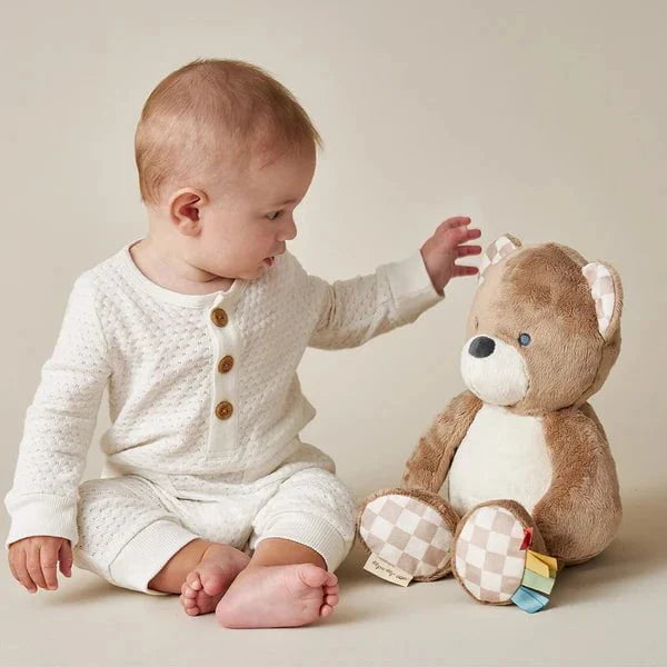 Itzy Ritzy Sweetie Snuggles Plush bear toy in tan with white belly as a baby reaches to pet its soft fur.