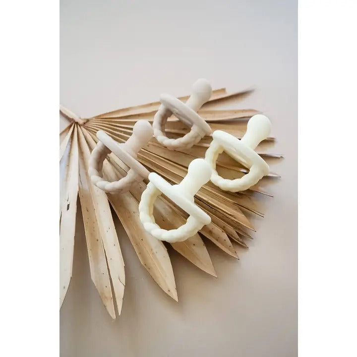 Itzy Ritzy Sweetie Soother Orthodontic Pacifier Sets on a dried palm frond, four white pacifiers showing orthodontic design.