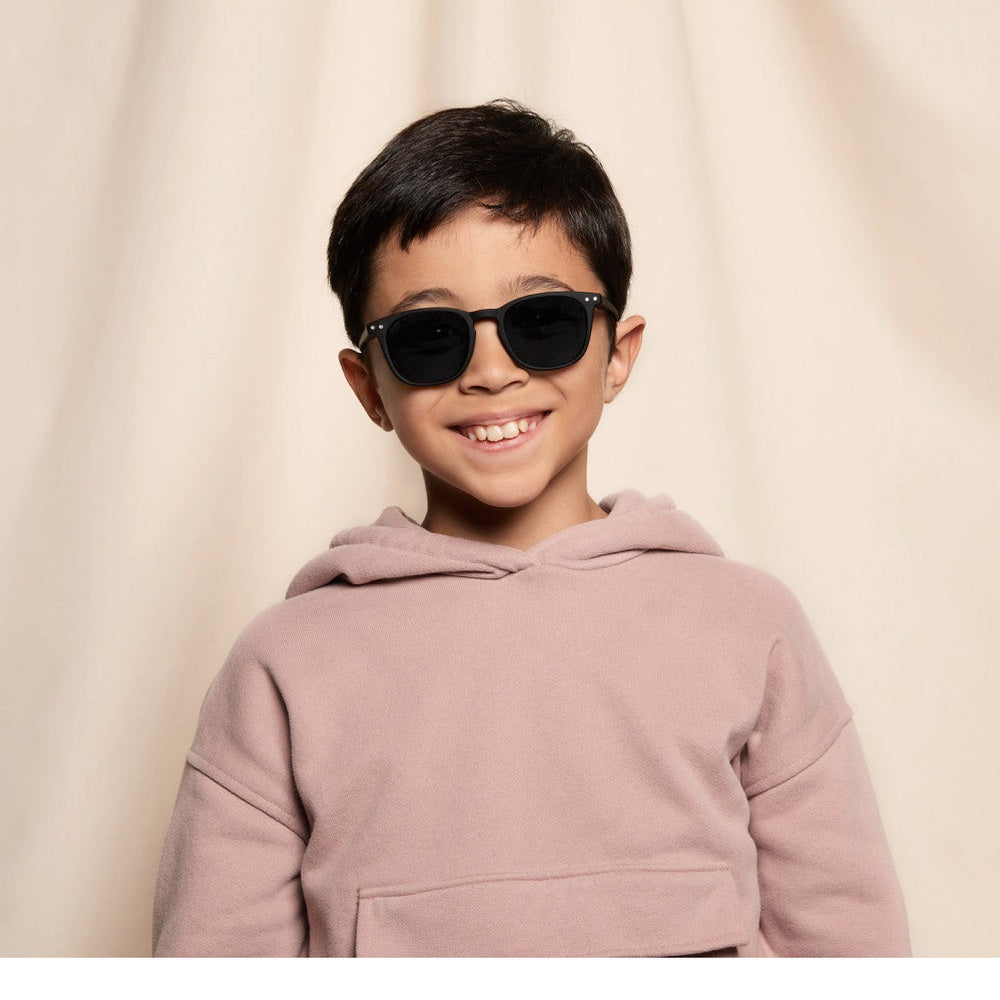 IZIPIZI Sun Junior #E Black sunglasses, black frames, worn by a smiling boy in a dusty pink hoodie