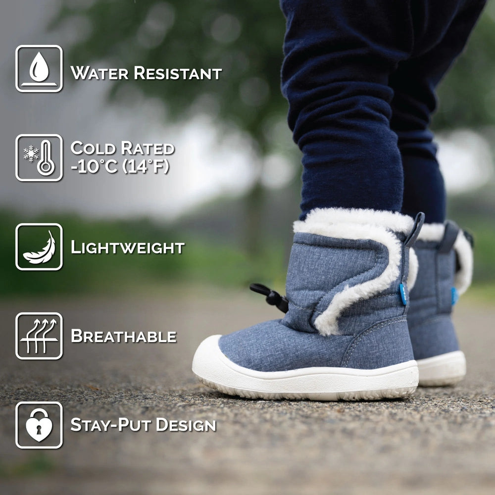 JAN & JUL Baby Winter Boots Bear in blue denim with white faux fur, water resistant and lightweight as a baby stands on the path.
