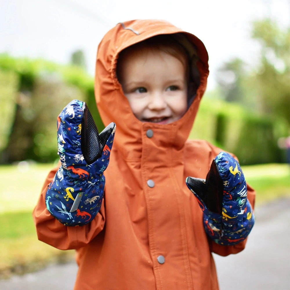 Jan & Jul Cozy Dry Mitten Space Dinos blue dinosaur print mittens worn by a smiling toddler raising hands in a rain jacket.