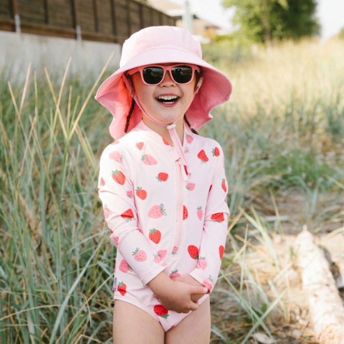 JAN & JUL Girls One Piece UV Swimsuit Pink Strawberry worn by a joyful child in a pink sun hat and sunglasses outdoors.