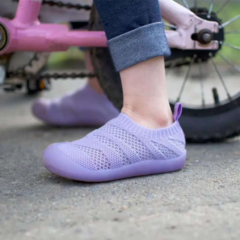 Jan & Jul Jelly Jumper Flow Shoes Lavender knit sock sneakers with pull tab on toddler's foot beside a pink bike.