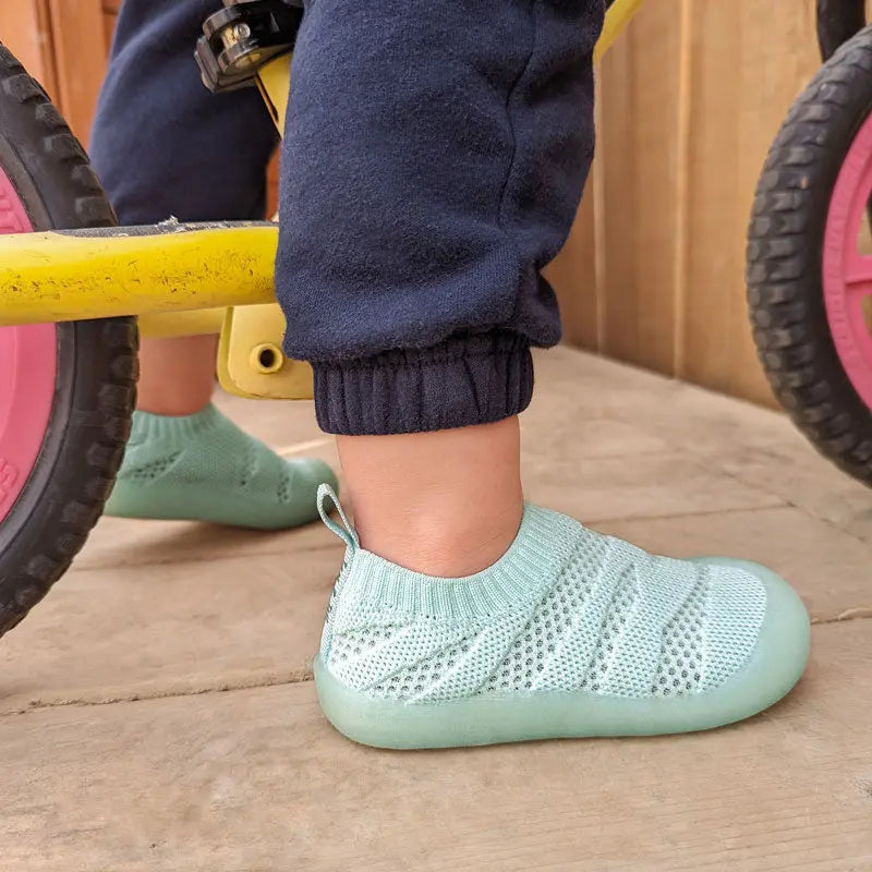 JAN & JUL Jelly Jumper Flow Shoes Mint knit sock-style toddler sneakers worn by a child beside a pink-wheeled bike.