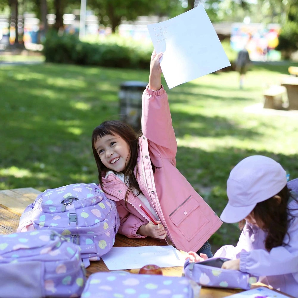 Jan & Jul Kids Backpack lavender with pastel hearts, open zipper pockets shown as kids at a park table.