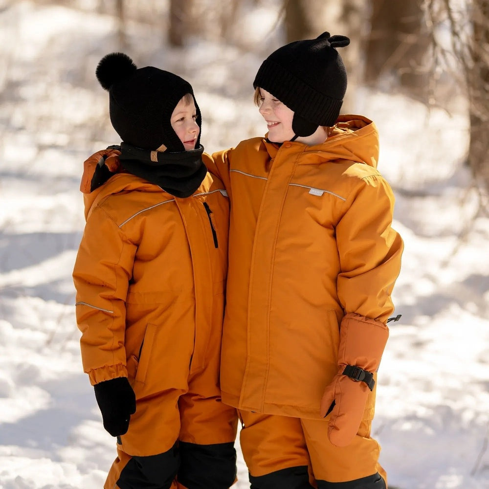 JAN & JUL Kids Bear Knit Hats Jet Black worn by two kids in orange snowsuits sharing a moment in a snowy forest.