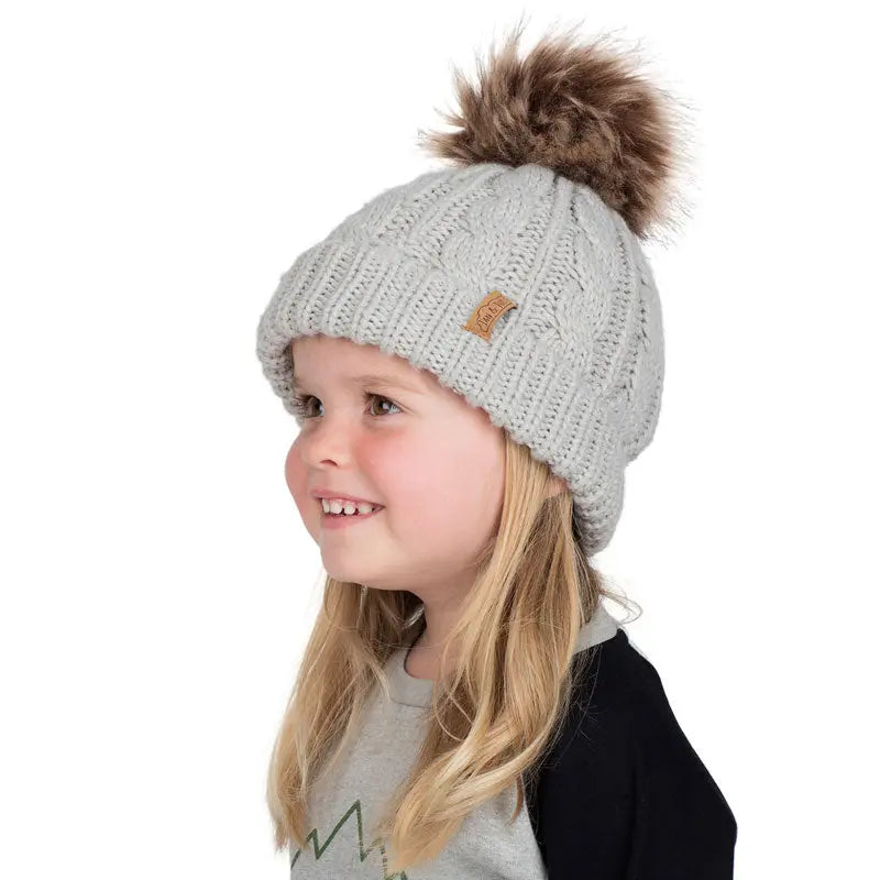 JAN & JUL Kids Cable Knit Twinning Beanie Grey worn by a smiling girl, showing cozy knit and fur pom-pom