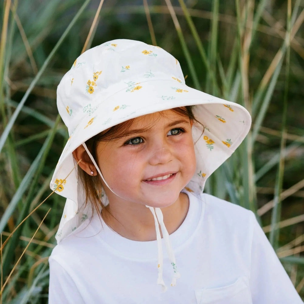 JAN & JUL Kids Cotton Adventure Hat, white with yellow flowers, chin ties, worn by a smiling girl outdoors.